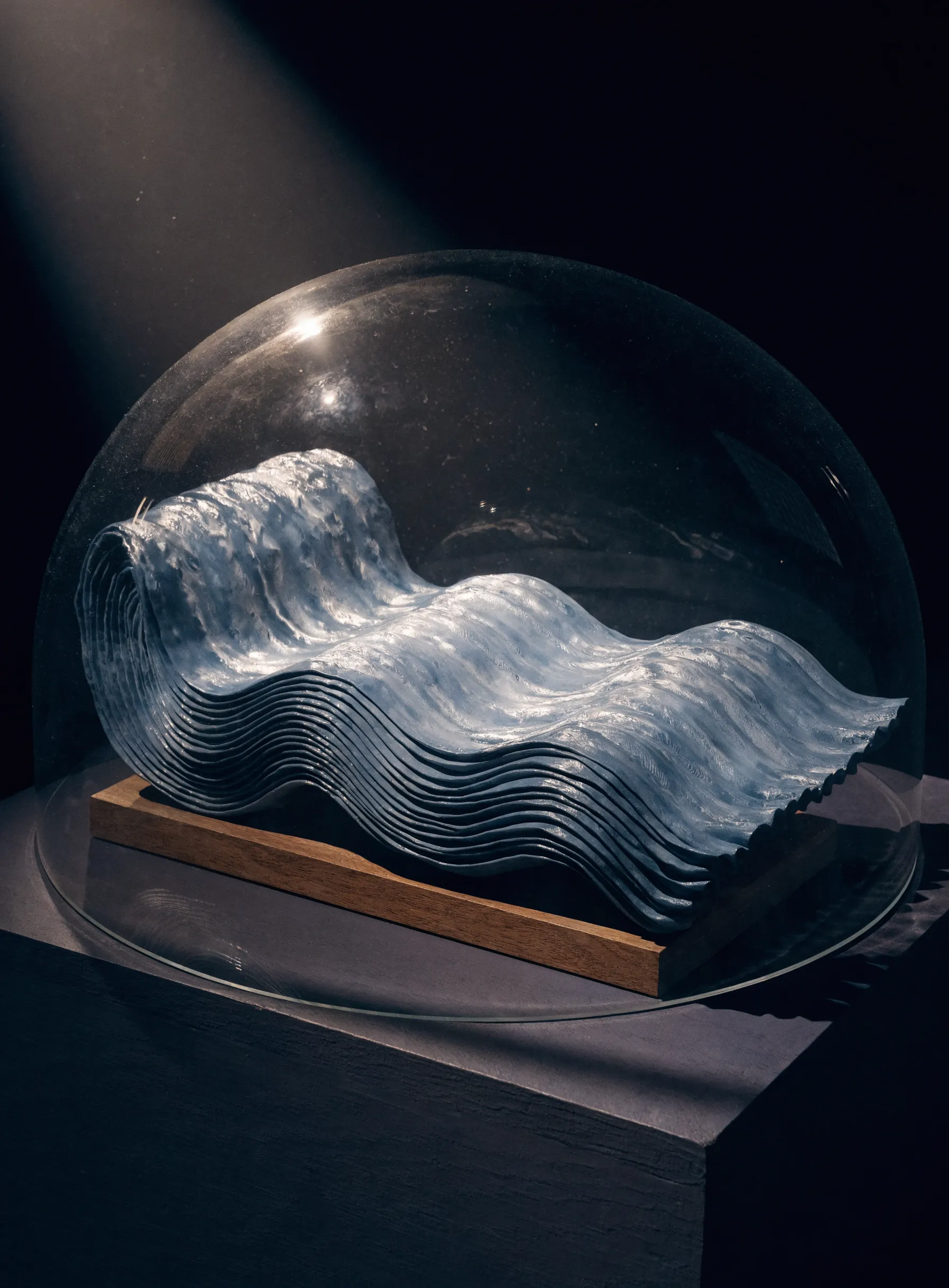 A river folded like pleated cloth, layered waves of pale water stacked under a round glass dome on a wooden base, one crest still curling mid-fold.